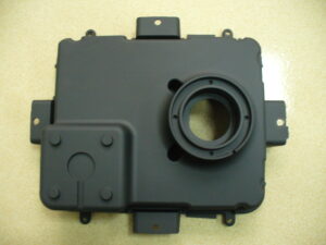 A close-up photograph of a matte black, machined metal optical housing with intricate internal light baffles, resting on a metal lab bench with electronic test equipment in the blurred background.